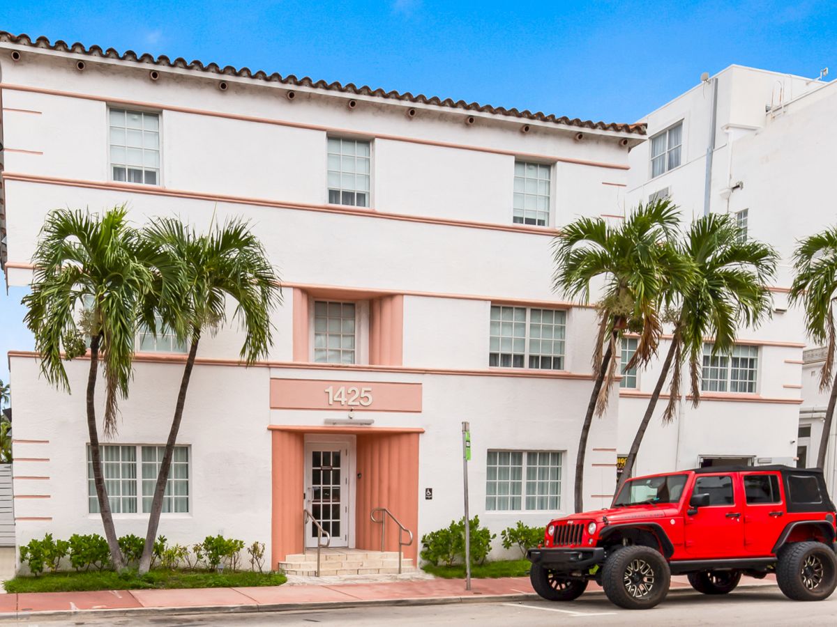 A three-story building with palm trees in front, featuring a red Jeep parked on the street.