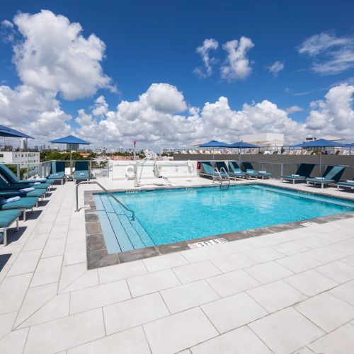 A rooftop swimming pool surrounded by loungers and umbrellas under a bright blue sky with scattered clouds.