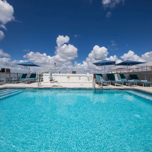 A rooftop pool with lounge chairs and umbrellas under a bright, partly cloudy sky.