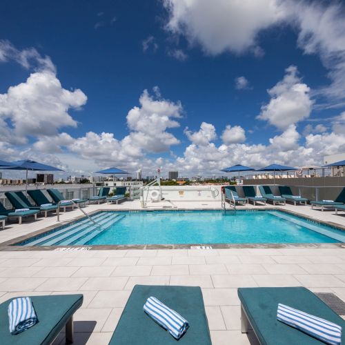 A rooftop swimming pool surrounded by sun loungers under a partly cloudy sky, with folded towels on the loungers.