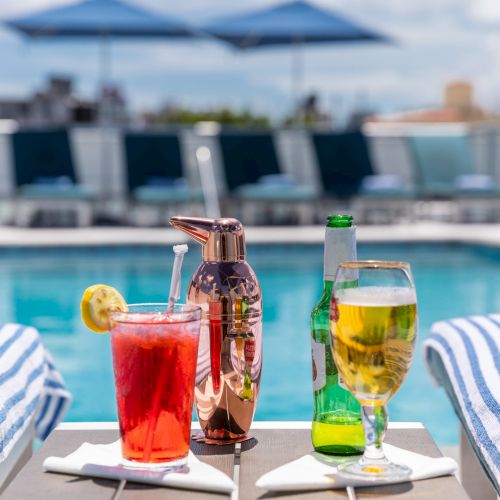 Drinks by a poolside, featuring a red cocktail with a lemon slice, a beer bottle, and wine glass, with striped towels in the background.