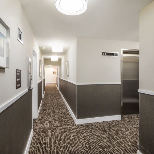 The image shows a hotel hallway with patterned carpet, framed art on walls, a silver elevator on the right, and bright ceiling lights.