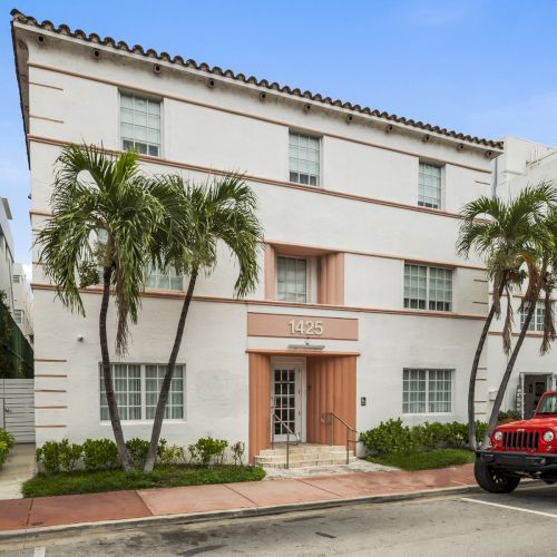 A three-story white building with palm trees in front, set on a street with a red vehicle parked nearby, under a clear blue sky.