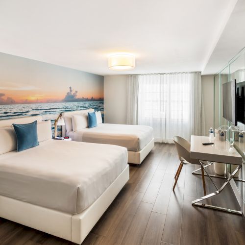 The image shows a modern hotel room with two beds, a wall mural of a sunset, a desk with chairs, and a TV mounted on a mirrored wall.