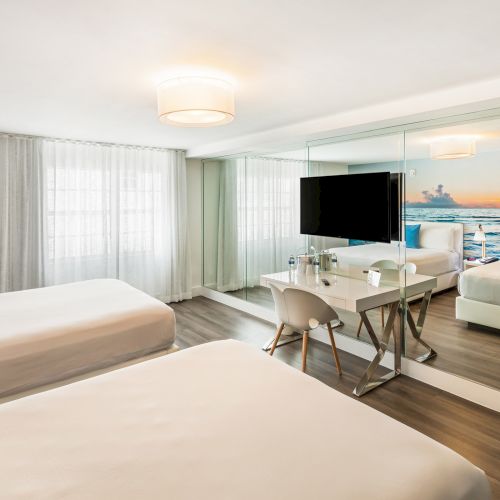 The image shows a modern hotel room with two beds, white linens, a desk, TV, chair, and a large mirror reflecting a beach scene.