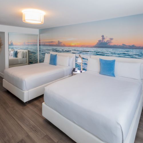 A modern hotel room with two beds, ocean-themed wall mural, white decor, blue pillows, wooden floor, and a desk with chairs.