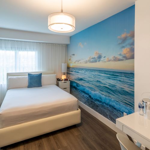 A modern bedroom with a large ocean mural, white bed, and desk. Light wood flooring and minimal decor create a serene atmosphere.