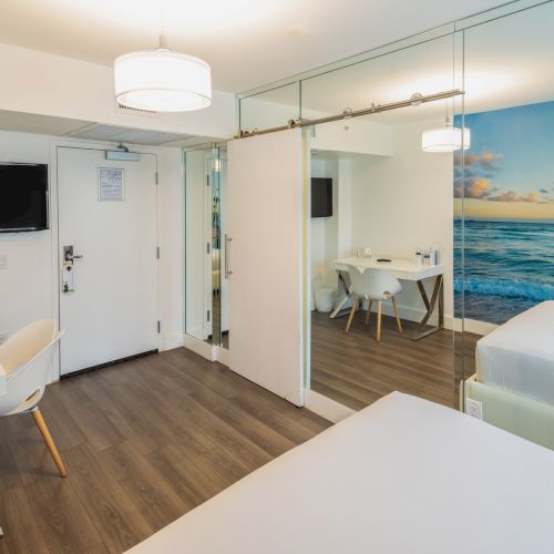 A modern hotel room with a double bed, wall art of the ocean, desk area, TV, and a small dining table, featuring a sleek design.