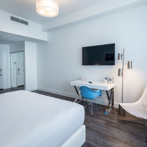 A modern hotel room with a bed, TV, desk, blue chair, and a white lounge chair on a wooden floor, all set against white walls.