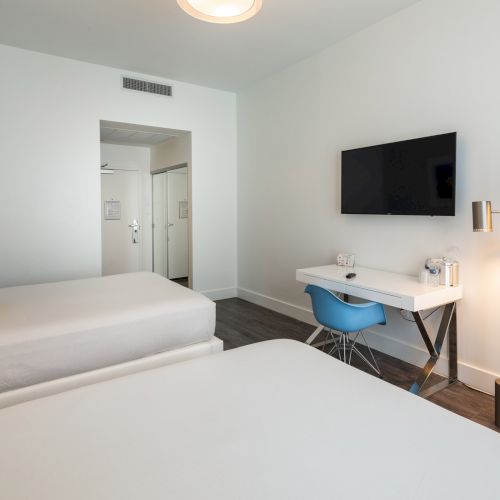 The image shows a modern hotel room with two beds, a wall-mounted TV, a desk with a blue chair, and a floor lamp.