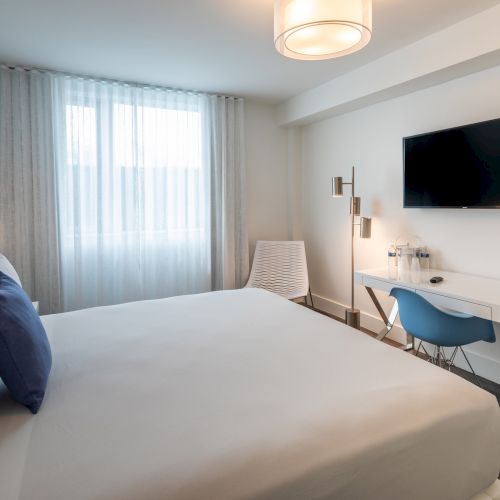 A modern hotel room with a large bed, blue chair, desk, TV, and window with sheer curtains.