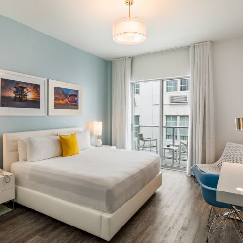 A modern hotel room with a bed, desk, TV, and balcony. It features blue and white decor with framed pictures on the wall.