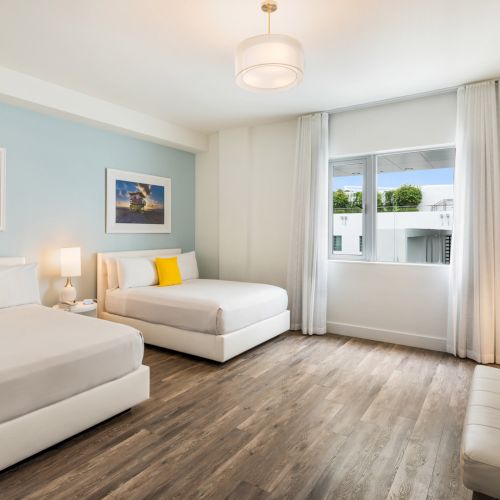 A modern bedroom with two beds, yellow cushions, a window with curtains, framed art on light blue walls, and wooden flooring.