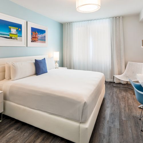 A modern, bright hotel room with a large bed, white furnishings, wall art, a desk with a blue chair, and wooden flooring.