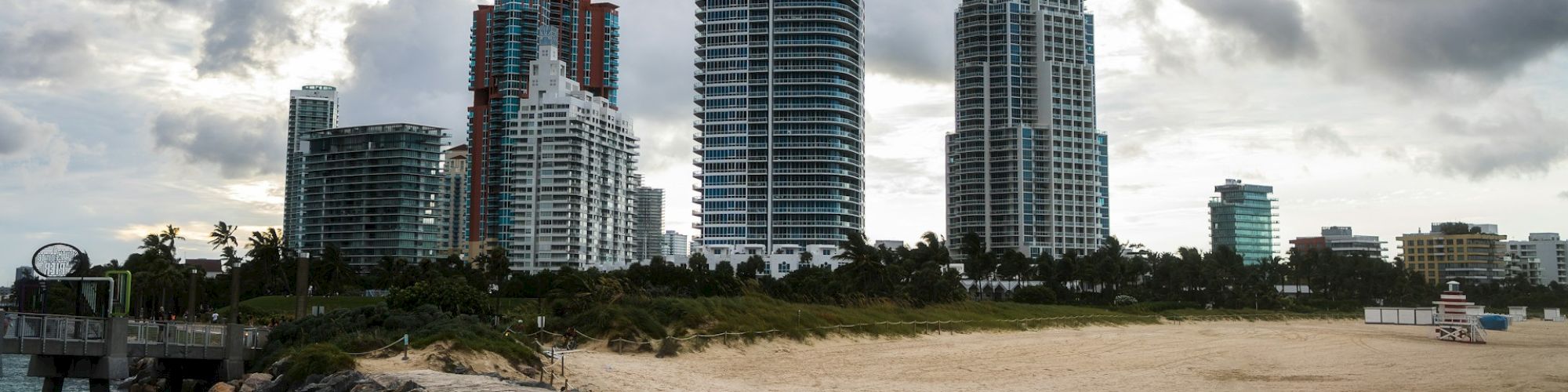 The image shows a rocky shoreline with a sandy area leading to a skyline of tall buildings under a partly cloudy sky.