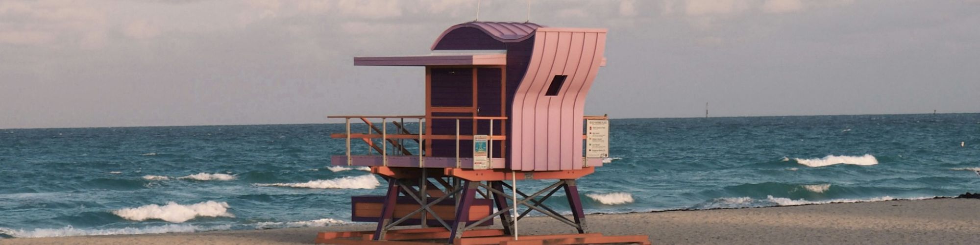 A colorful lifeguard tower stands on a sandy beach with the ocean and sky in the background, under a partly cloudy sky.