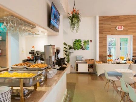 The image shows a well-lit dining area with a buffet setup including pastries and hot food next to a seating area with modern decor and plants.