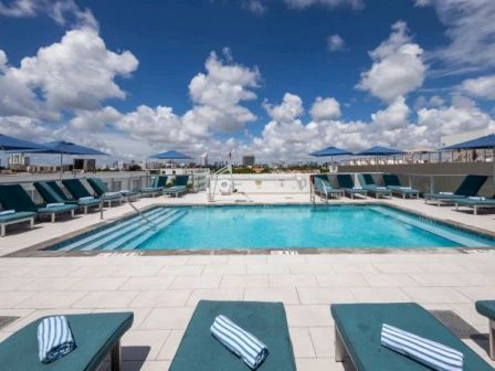 This image shows a rooftop pool area with lounge chairs, blue towels, and umbrellas under a partly cloudy sky.