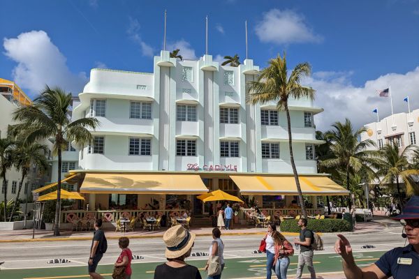 People walking on a sidewalk in front of a multi-story Art Deco building with yellow awnings, palm trees, and a clear blue sky.