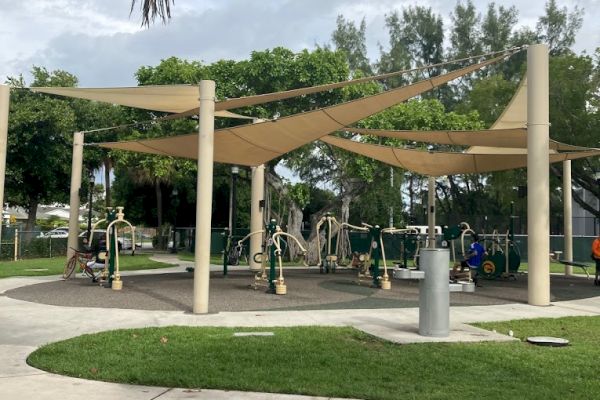 The image shows an outdoor fitness area with exercise equipment, covered by shade structures, surrounded by grass, trees, and a paved walkway.