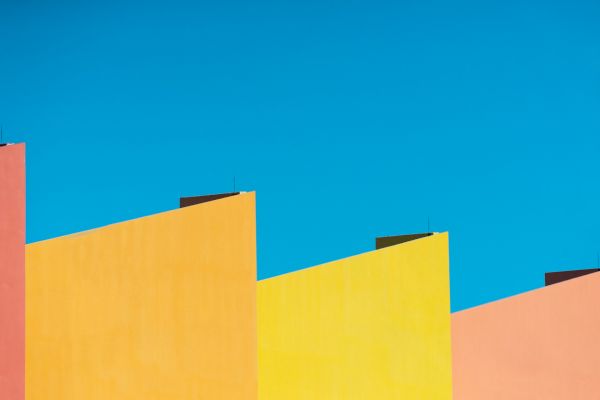 The image shows colorful, geometric building rooftops with vibrant shades of pink, orange, and yellow against a clear blue sky.