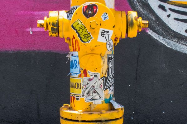 The image shows a bright yellow fire hydrant covered in various colorful stickers and graffiti, with a mural painted on the wall behind it.