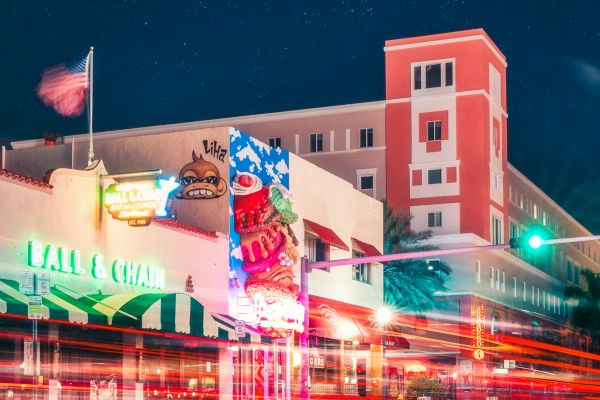 A vibrant night street scene with colorful buildings, neon lights, and light trails from passing vehicles. The sky is filled with stars.