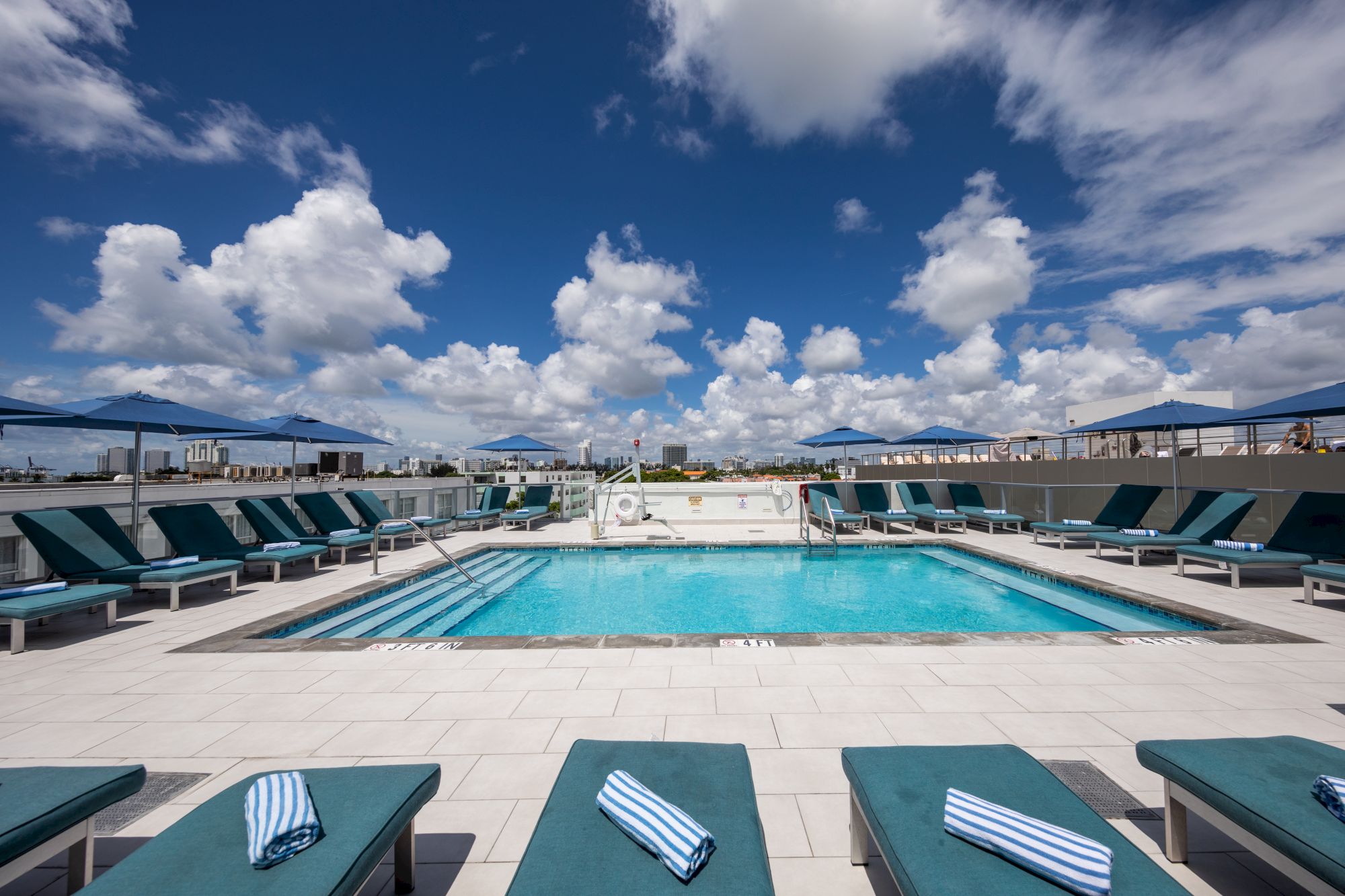 A rooftop pool area with turquoise loungers, neatly placed towels, blue umbrellas, and a backdrop of a partly cloudy sky .