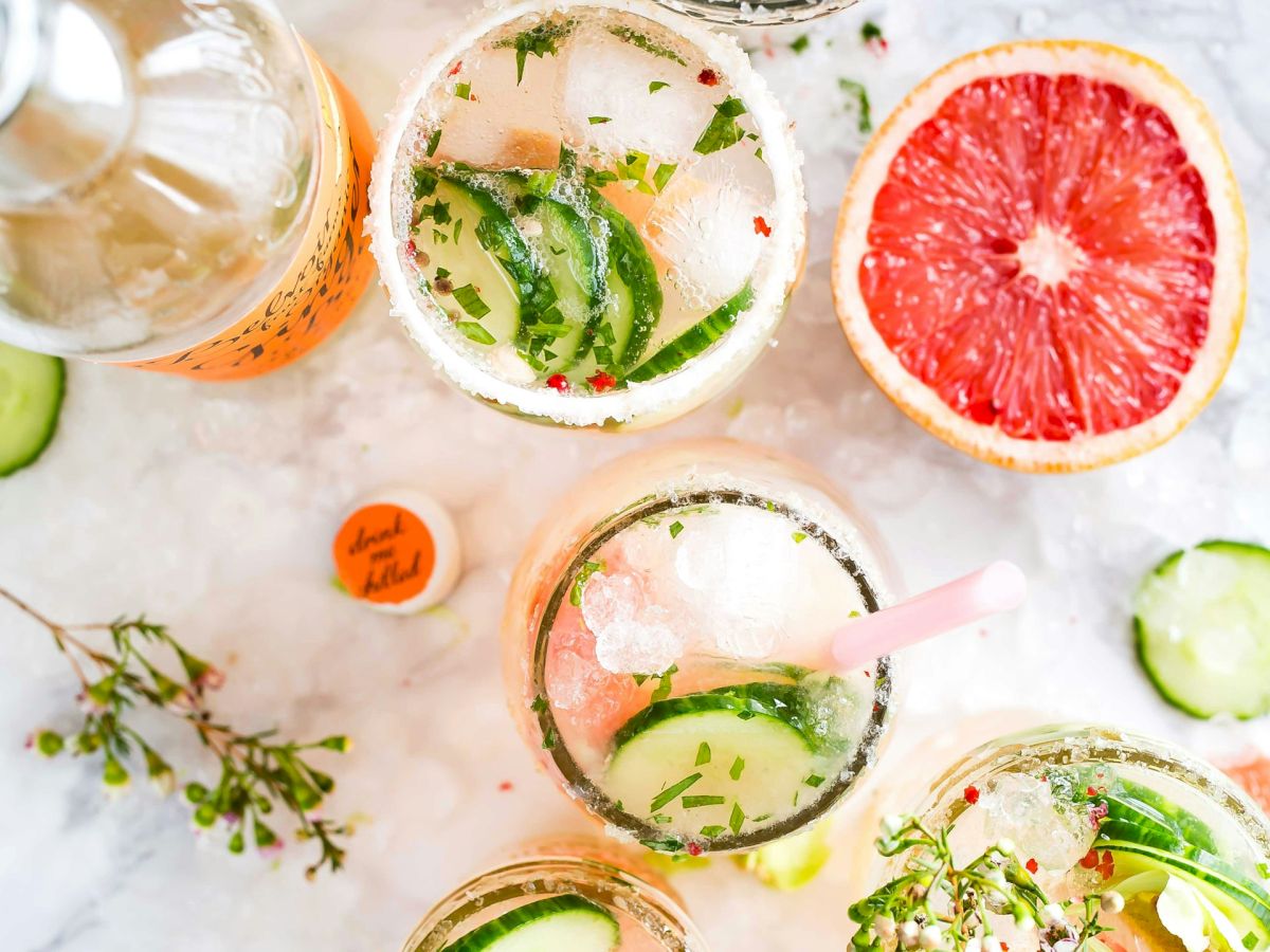 The image shows fruit-infused drinks with herbs and garnishes, a grapefruit half, ice, a bottle, and cucumber slices on a marble surface.